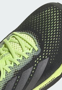 Zapatilla de running negra con parte superior de malla y acentos en verde neón, superficie texturizada y un diseño estructurado. Incluye cordones visibles y logo.