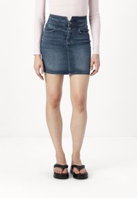Femme portant une mini-jupe en jean bleu taille haute, un haut pâle à manches longues et des sandales compensées noires, debout sur un fond blanc uni.