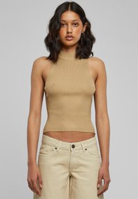 Kvinde med skulderlangt mørkt hår iført en tætsiddende beige ærmeløs ribbet top og lyse beige bukser, stående foran en ensfarvet grå baggrund.
