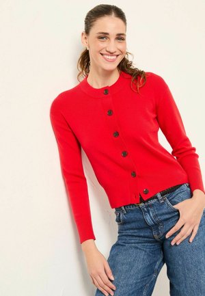 Red cardigan made of knit fabric, featuring a round neckline, long sleeves, and black buttons down the front. Worn over blue jeans.
