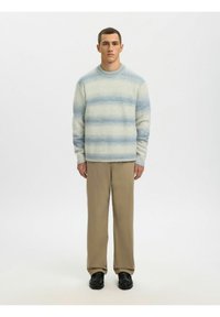 Homme debout portant un pull rayé bleu clair et blanc, un pantalon beige et des chaussures noires, sur un fond blanc uni.