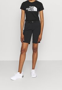 Černé tričko s krátkým rukávem a bílým logem, spárované s černými šortkami a bílými teniskami. Oblečení má neformální, sportovní design.