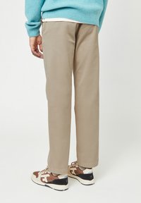 Persona che indossa pantaloni beige, un maglione turchese e sneaker marroni, crema e nere, in piedi con la schiena rivolta verso la fotocamera.