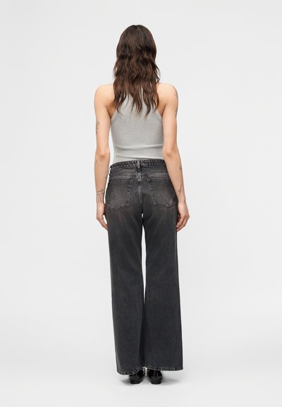 Γυναίκα με μακριά κυματιστά μαλλιά που φοράει γκρι αμάνικο τοπ και μαύρο τζιν ψηλόμεσο με φαρδιά μπατζάκια, στέκεται μπροστά σε απλό φόντο.