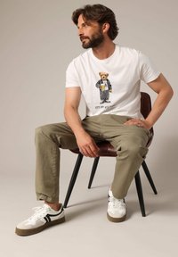 Witte t-shirt met berenprint, beige broek en witte sneakers met zwarte accenten, zittend in een stoel in mid-century stijl.
