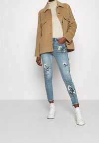 Ruskea villasekoitejakku, jossa kaksi etutaskua, beige pooloneule, kulutetut vaaleansiniset farkut, joissa kukkaistuja kirjailuja, ja valkoiset tennarit.