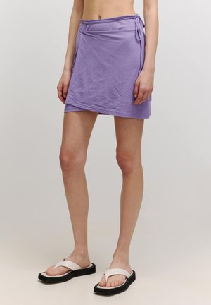 Paarse mini rok met een wikkelontwerp, gemaakt van zachte stof, met een strik taille detail en een asymmetrische onderrand. Gecombineerd met witte sandalen.