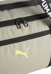 Bolso de lona gris con cierres de cremallera negros, que presenta un logo de Puma amarillo y asas negras, mostrando una textura ribeteada vertical.
