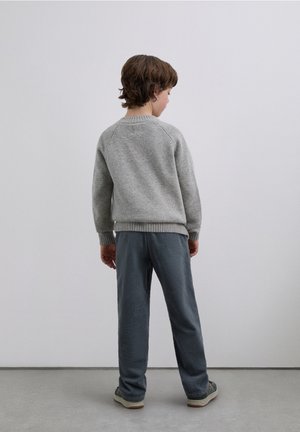 Pull gris clair en maille avec poignets et ourlet côtelés, associé à un pantalon gris foncé texturé. La personne est vue de dos.