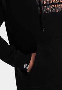 Felpa nera con cappuccio e tasca a marsupio frontale, caratterizzata da una grafica testurizzata in arancio e nero. Polsini a costine e un piccolo logo sulla manica.