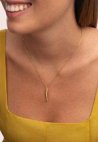 Collana con pendente a forma di pesce rosso su una sottile catena, indossata su un collo nudo. La figura è dettagliata con squame, su un fondo di tessuto giallo.