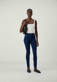 Γυναίκα φορώντας λευκή αμάνικη μπλούζα, μπλε στενά τζιν, μαύρες στιλιζαρισμένες πλατφόρμες, κρατώντας μια μαύρη τσάντα ώμου σε απλή φόντο.