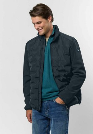 Homme souriant portant une veste matelassée noire par-dessus un pull zippé bleu sarcelle et un jean bleu, une main dans sa poche.