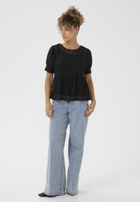 Top in pizzo nero con maniche a sbuffo corte e orlo svasato, abbinato a jeans a gamba larga blu chiaro e scarpe nere con punta affilata.