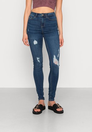 Frau trägt hoch taillierte, ausgewaschene, enge blaue Jeans, ein kurz geschnittenes bordeauxrotes Top und schwarze Slip-On-Sandalen mit goldenen Schnallen.