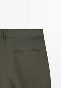 Dark olive green pants showing the waistband, belt loops, and a single welt back pocket on a plain background.