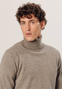 Lichtbruine coltrui van zacht breigoed, met een hoge kraag en geribbelde details op de hals, schouders en zoom.