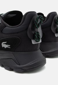 Sorte sneakers med tekstureret stof, udstyret med grønne spottede snørebånd og en gummisål. Lacoste-logo på siden, minimalistisk design.
