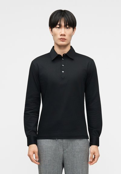 Hombre de pie con una camisa negra de manga larga con cuello y botones, y pantalones grises con rayas finas, sobre un fondo neutro.