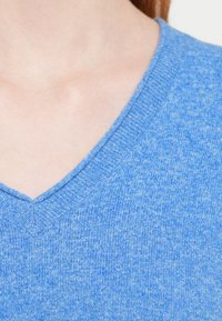 Zils adījuma džemperis ar V-veida izgriezumu. Tam ir mīksta tekstūra un viegli raibs raksts, kas uzlabo tā kopējo izskatu.