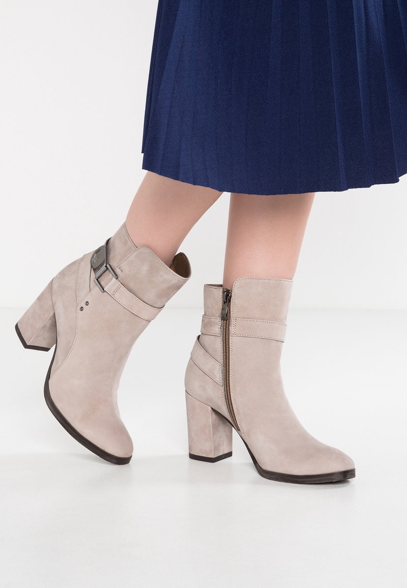 Bottines en daim beige clair à talons blocs, fermeture zippée latérale et boucle décorative sur la lanière, portées avec une jupe plissée bleu marine.