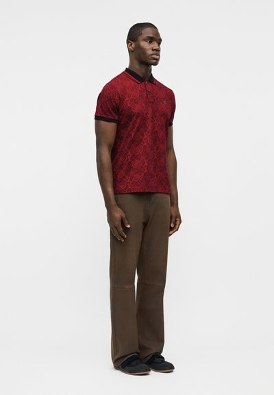 Hombre de pie mirando hacia adelante, vestido con una camisa tipo polo roja con estampado, pantalones marrones y zapatos negros, sobre un fondo blanco liso.