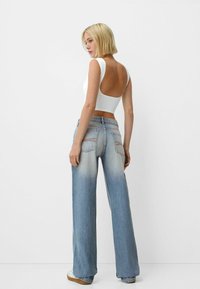 Lyseblå bredbenede jeans med en falmende gradient, parret med en hvid ærmeløs top med lav rygdesign. Flade, minimalistiske sko.