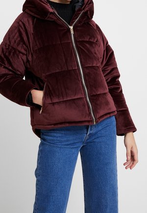 Henkilö, jolla on päällään burgundinpunainen samettitoppatakki, jossa on huppu, vetoketju edessä ja sivutaskut. Hänellä on myös siniset farkut ja musta villapaita.