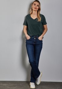 Tmavě zelené tričko s výstřihem do V, modré rozšířené džíny a bílé tenisky. Hladká tkanina bez vzorů a akcentů.