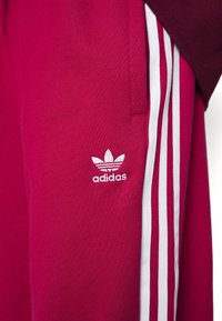 Maroon joggerbyxor med vita tre-stripes detaljer längs sidorna och en broderad Adidas-logga på nedre vänstra benet.