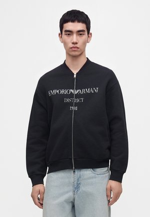 Jeune homme portant un sweat zippé noir avec le texte "Emporio Armani District 1981", associé à un jean bleu clair, sur un fond uni.