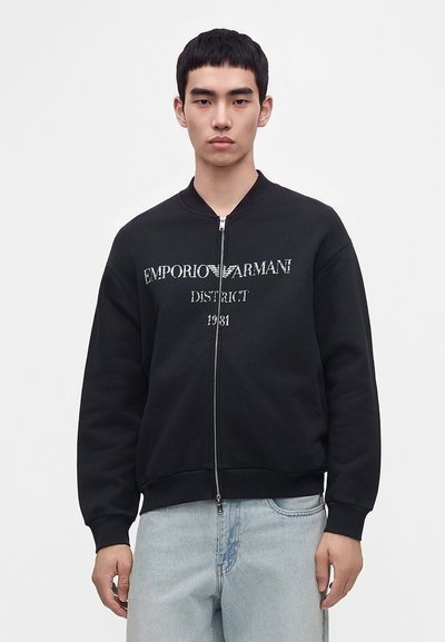 Jeune homme portant un sweat zippé noir avec le texte "Emporio Armani District 1981", associé à un jean bleu clair, sur un fond uni.