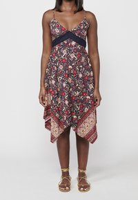 Robe à motifs floraux dans des teintes marine, rouge et crème ; bretelles spaghetti ajustables ; ourlet asymétrique ; accent en dentelle sous la poitrine ; associée à des sandales dorées.