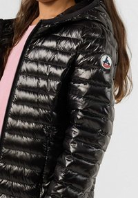 Chica con una chaqueta acolchada negra brillante y capucha, con un parche de logo rojo y blanco en la manga, sobre una blusa de color rosa claro.