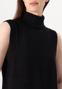Μαύρο χωρίς μανίκια πουλόβερ με ψηλό λαιμό, φτιαγμένο από μαλακό ύφασμα με ραβδωτές λεπτομέρειες στον λαιμό και στα άκρα των μανικιών, που παρουσιάζει ομαλή υφή.