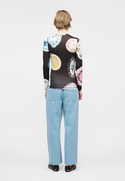 Άτομο με κοντά ανοιχτόχρωμα μαλλιά, ντυμένο με μακρυμάνικη μπλούζα με πολύχρωμα σχέδια από πιάτα και ανοιχτό μπλε τζιν παντελόνι με φαρδιά ζιπ κιλότ, στέκεται με την πλάτη προς τα πίσω.