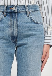 Ανοιχτό μπλε τζιν παντελόνι με ψηλή μέση, πέντε τσέπες και αντίθεση ραφών. Το ύφασμα έχει ελαφριά υφή και εμφάνιση ξεθωριασμένου πλυσίματος.