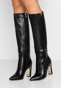 Svarta läderstövlar, höga till knäna, med spetsig tå och en klack i blockform med guldaccenter. Har en sidodragkedja för enkel påtagning.