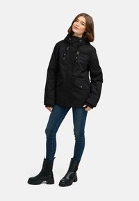 Svart isolerad jacka med dragkedja, justerbar huva och två framfickor; kombinerad med blå jeans och svarta ankelstövlar.