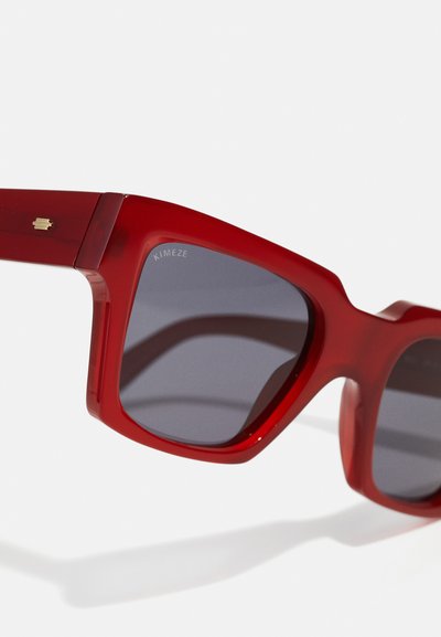 Ochelari de soare roșii cu rame groase și lucioase. Lentile întunecate cu branding minimal în colțul stâng sus al ramei. Design unghiular.