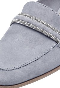 Mocassin en daim bleu clair avec deux bandes argentées ornées de strass, texture lisse, bout rond, et semelle contrastante couleur noisette.
