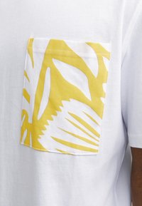 Hvid t-shirt med en gul lomme med et hvidt bladmønster. Lommen er syet pænt fast til fronten af trøjen.