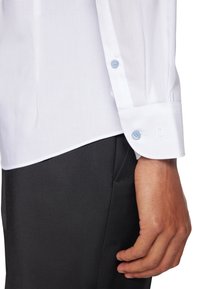 Nahaufnahme eines Arms einer Person, die ein weißes Hemd mit hellblauen Knöpfen und schwarze Hose trägt, Hand entspannt an der Seite.