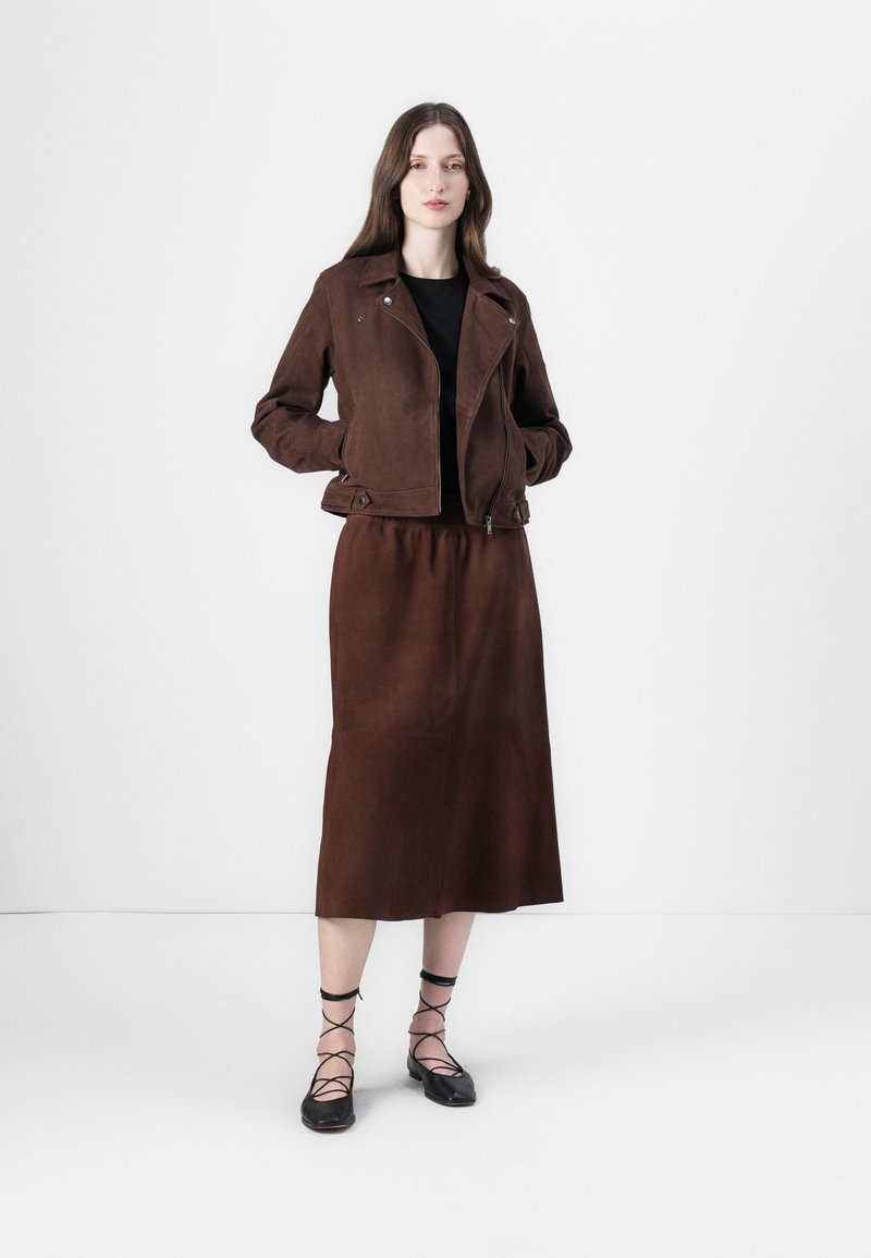 Brązowa skórzana kurtka z zamkiem błyskawicznym, noszona na czarnej bluzce. Pasująca brązowa spódnica sięgająca do połowy łydki. Czarne sznurowane buty dopełniają stylizację.