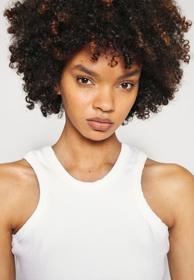 White ribbed tank top with wide shoulder straps, featuring a close neckline. Skin tone is warm, with curly hair showing hints of brown.