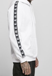 Uomo che indossa una felpa bianca con nastro logo nero sulle maniche e pantaloni neri, mostrando una mano tatuata e un braccialetto al polso.