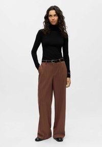 Pull à col roulé noir associé à un pantalon large à rayures marron. Ceinture à la taille, texture lisse et chaussures noires unies. Fond uni.