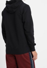 Černá bavlněná mikina s kapucí, dlouhými rukávy a žebrovanými manžetami. Viditelné švy a volný střih. Nošeno s vínovými kalhotami.