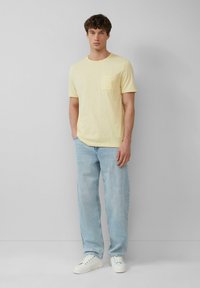 Lichtgele T-shirt met ronde hals en een borstzak. Gecombineerd met lichtblauwe rechtmodel jeans en witte sneakers, tegen een neutrale achtergrond.