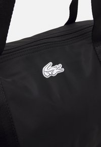 Sac en tissu noir avec une fermeture éclair sur le dessus et un logo crocodile blanc cousu sur le devant, avec deux bandoulières noires.